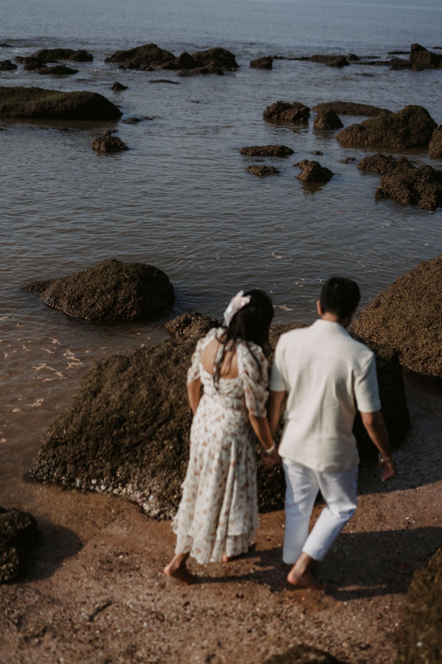 Two figures sitting by a rocky coastline at sunset with ocean waves washing against the shore.
