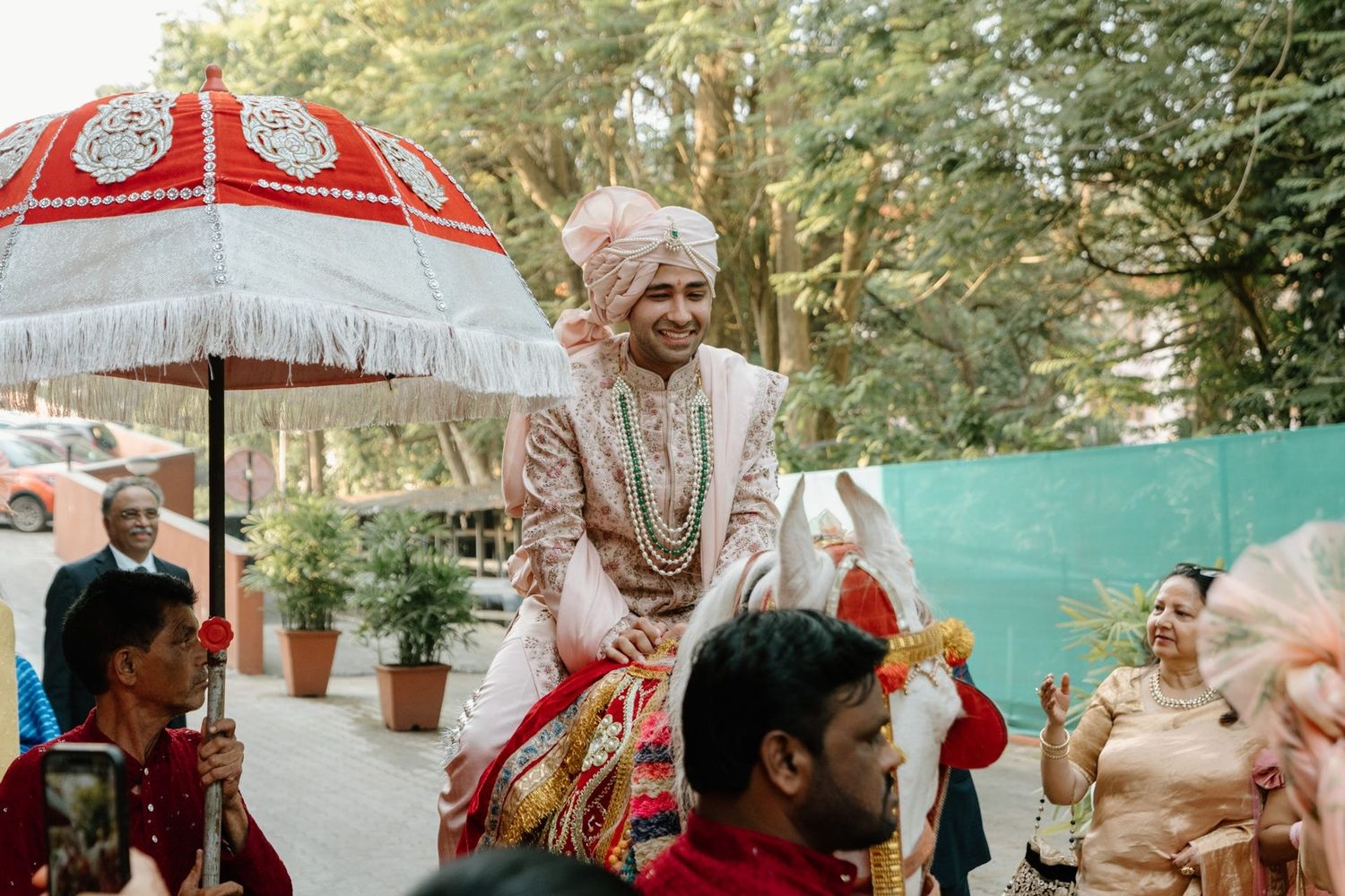 A traditional Indian wedding procession with a decorative red umbrella and ornate ceremonial attire in an outdoor garden setting.