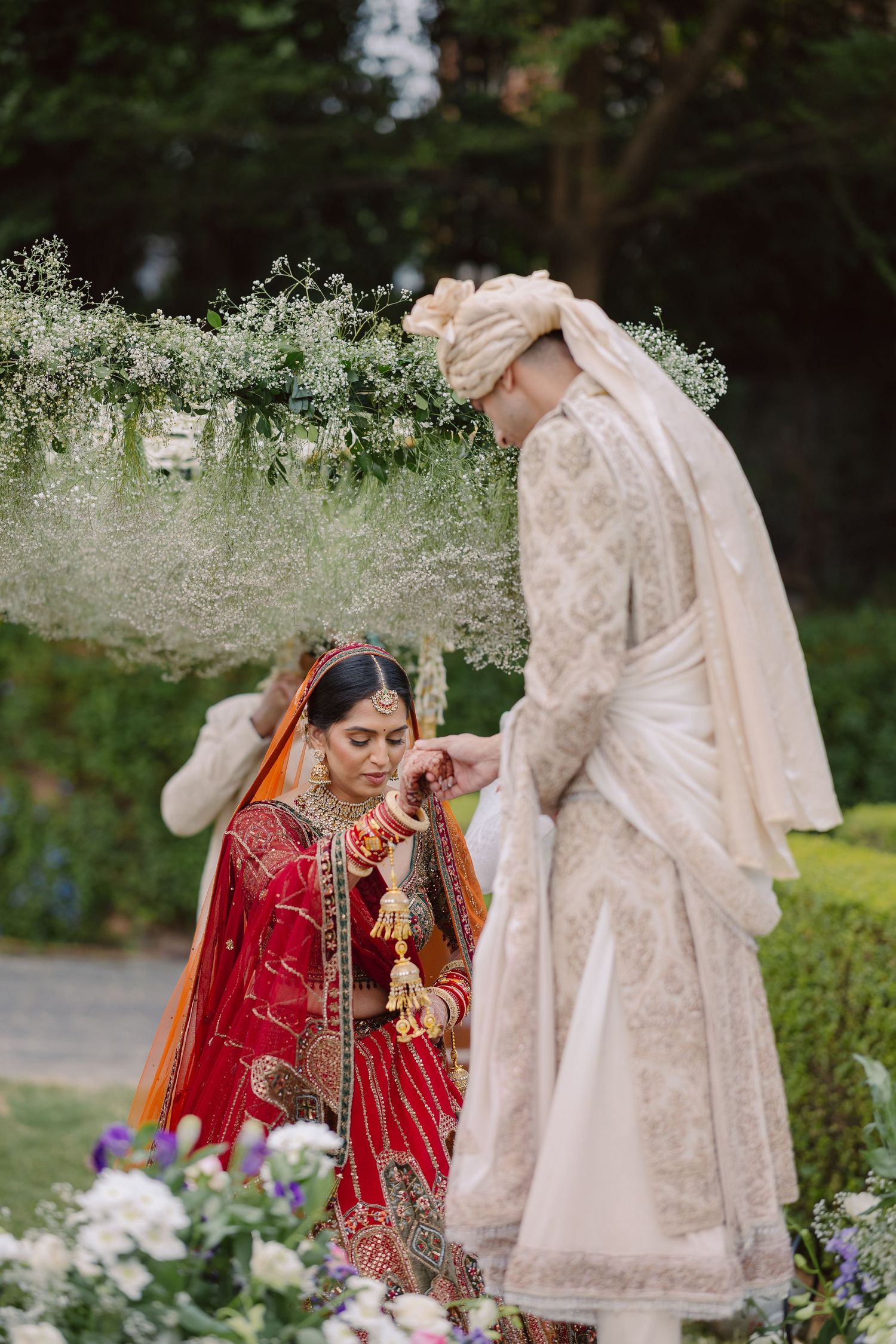 Traditional Indian wedding scene with bride in red lehenga and guests in cream outfits in garden setting.