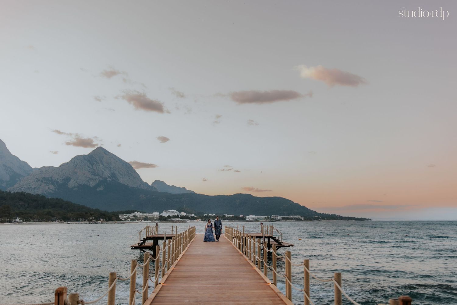 Couple stands on a wooden pier at sunset with mountain backdrop and calm sea in a romantic coastal scene.