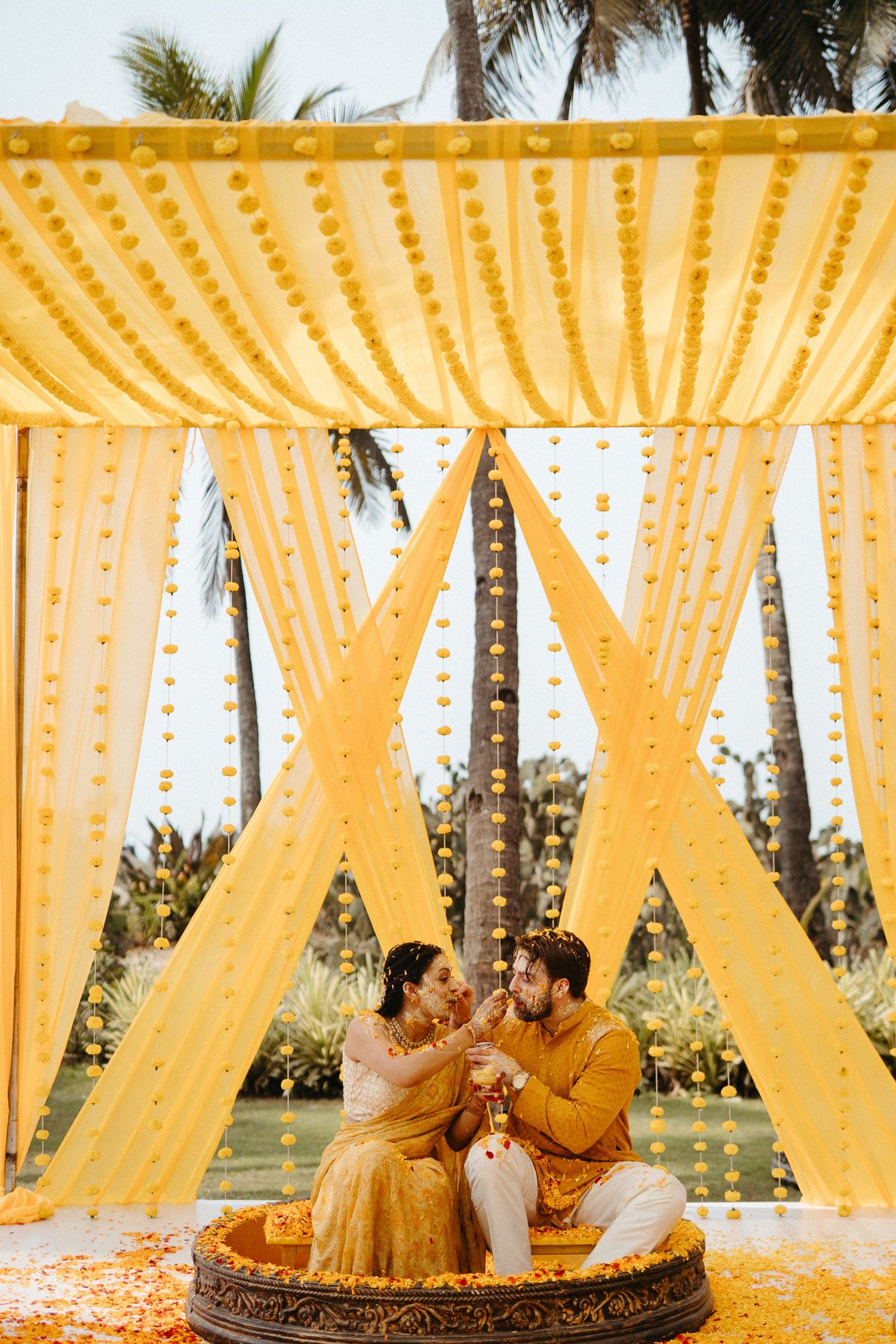 Couple in yellow outfits sitting under golden marigold mandap during traditional Indian haldi ceremony.