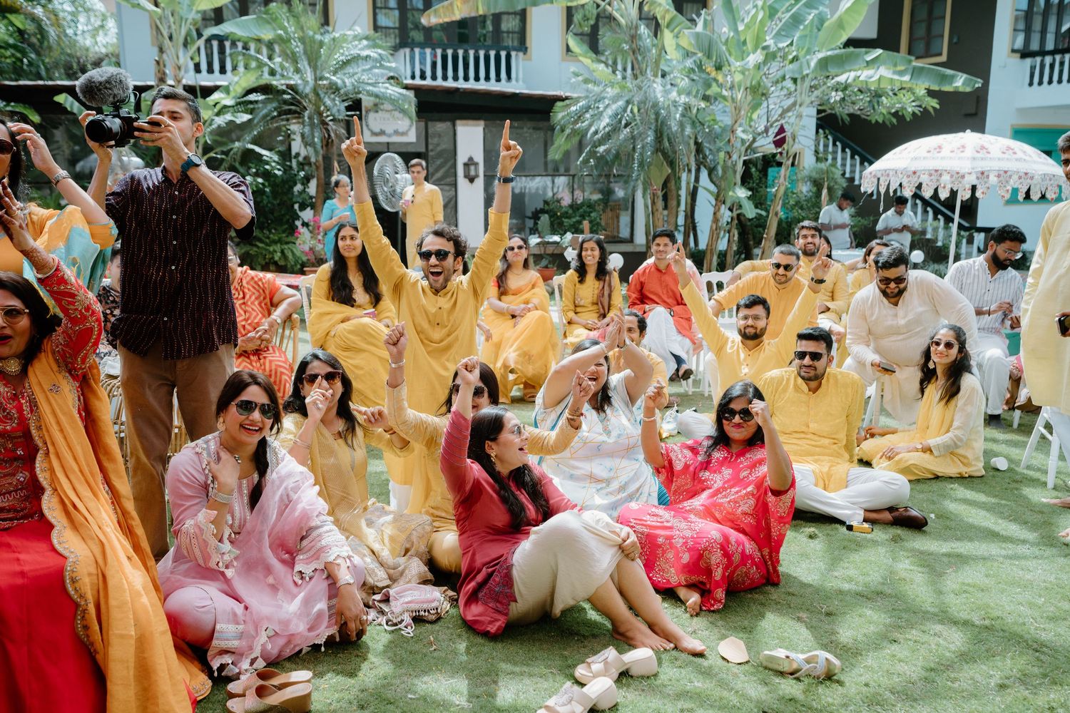 Joyful Indian wedding guests in yellow and pink outfits celebrate outdoors, raising hands in festive cheer.