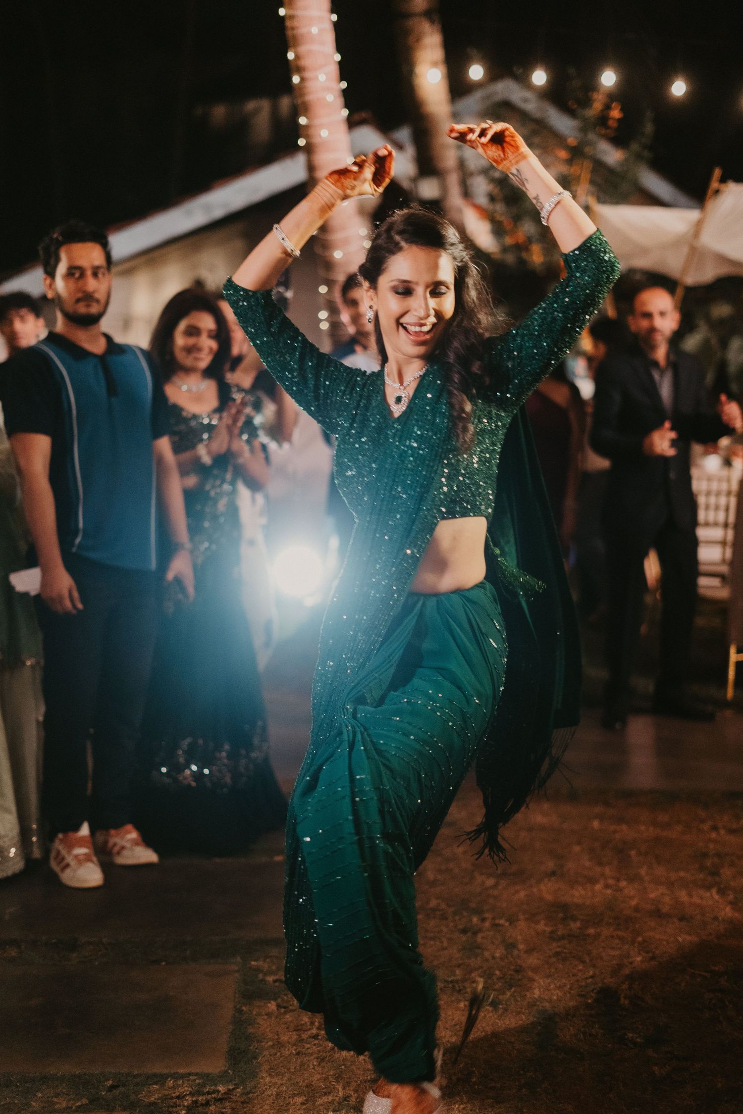 Woman in green sequin lehenga dancing joyfully at an outdoor Indian wedding celebration at night.
