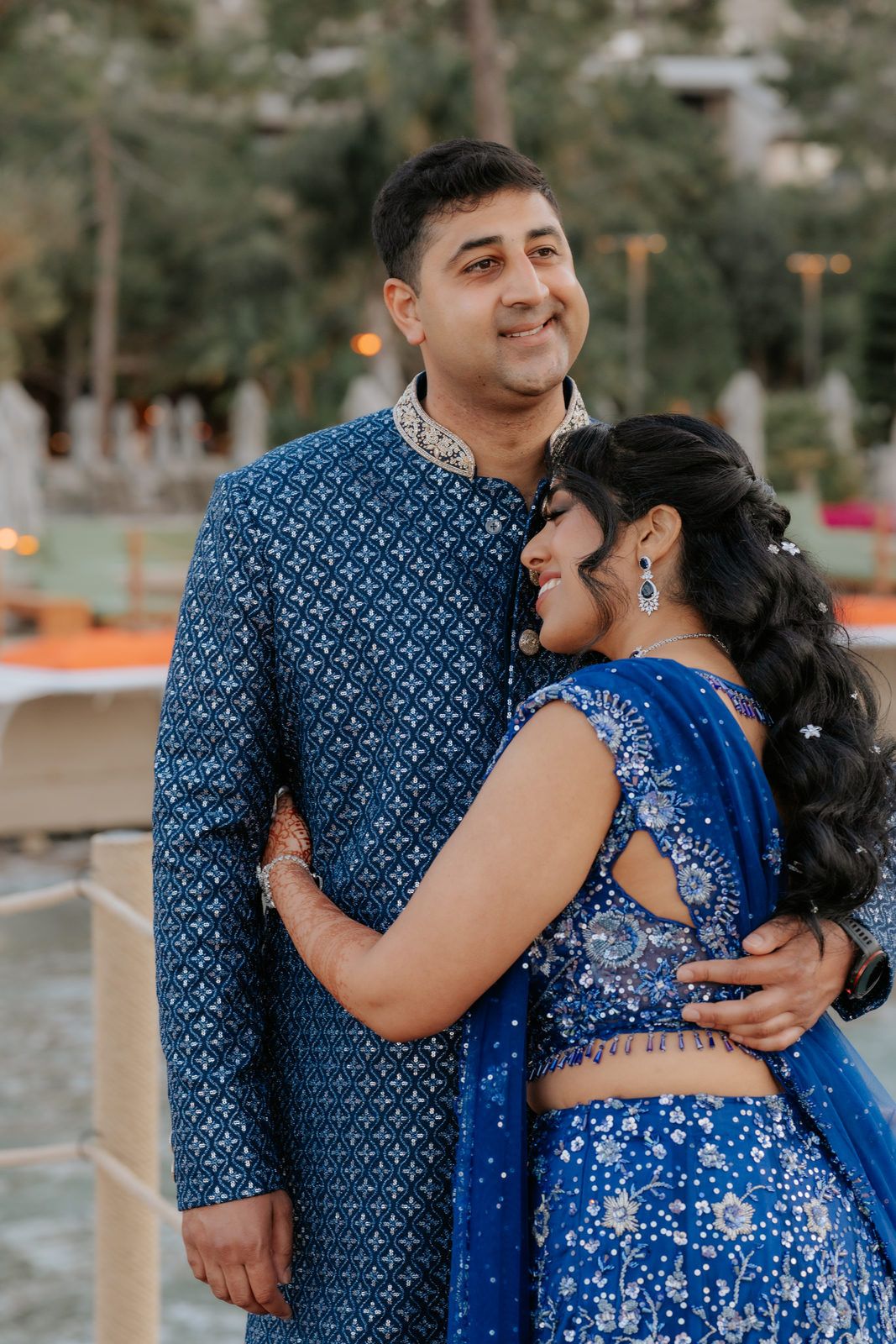 Couple in matching royal blue Indian wedding attire embrace outdoors, man in sherwani and woman in embroidered lehenga.
