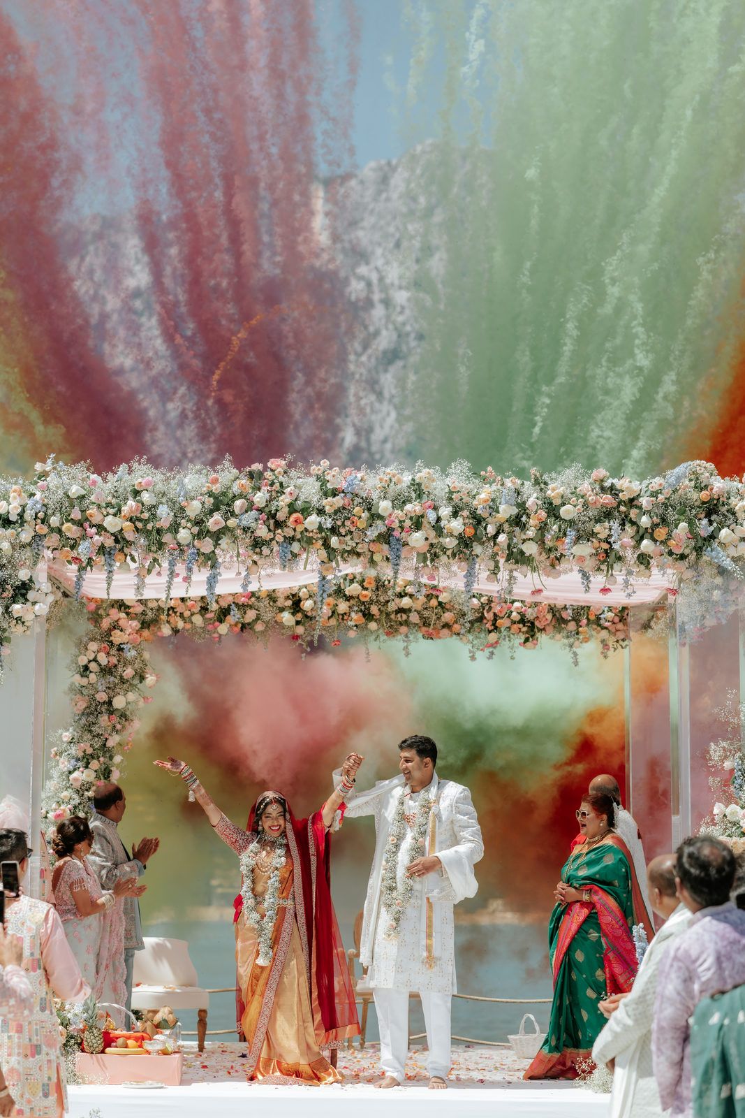 Indian wedding couple celebrates under a floral mandap with colorful powder clouds during ceremony.