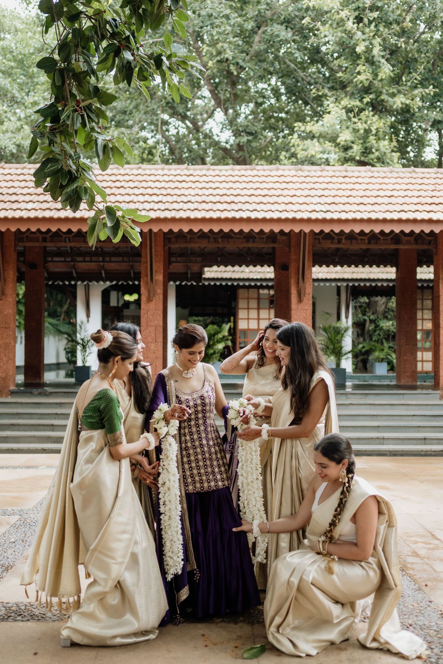 Five women in elegant cream silk sarees gather outdoors at a traditional Indian wedding venue surrounded by lush greenery.
