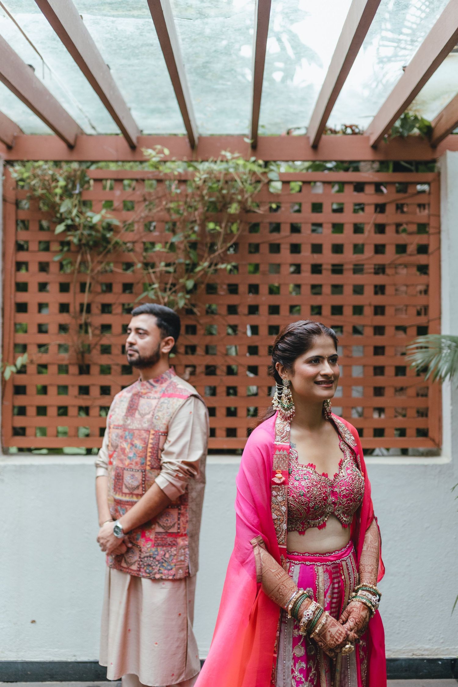 Indian bride in pink lehenga and groom in cream kurta pose separately at an outdoor wedding venue.