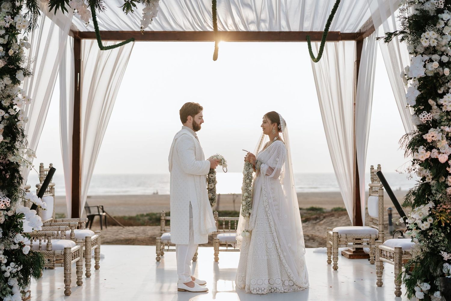 Couple in white attire exchanging vows under a floral arch at a beachside wedding ceremony.