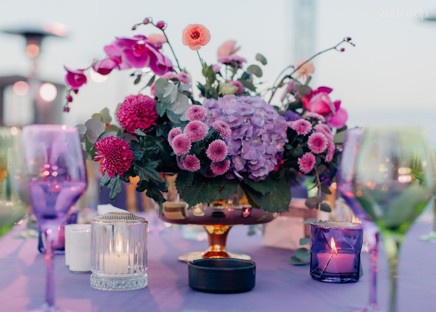 Elegant floral centerpiece with purple hydrangeas and pink dahlias on a lavender table with candles and glassware.