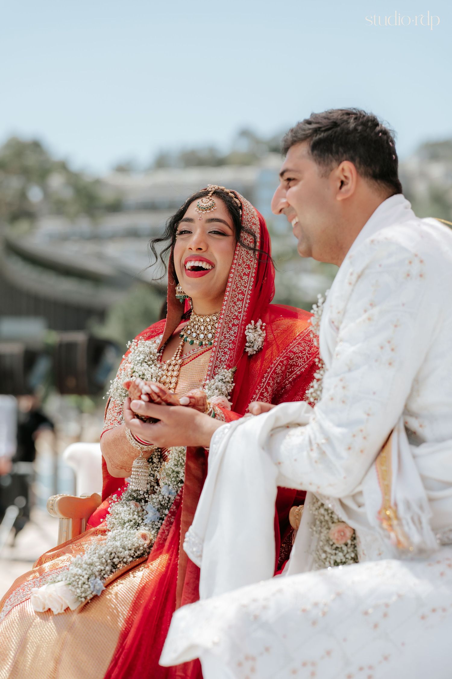 Bride in red lehenga and groom in white sherwani laughing joyfully during traditional Indian wedding ceremony.