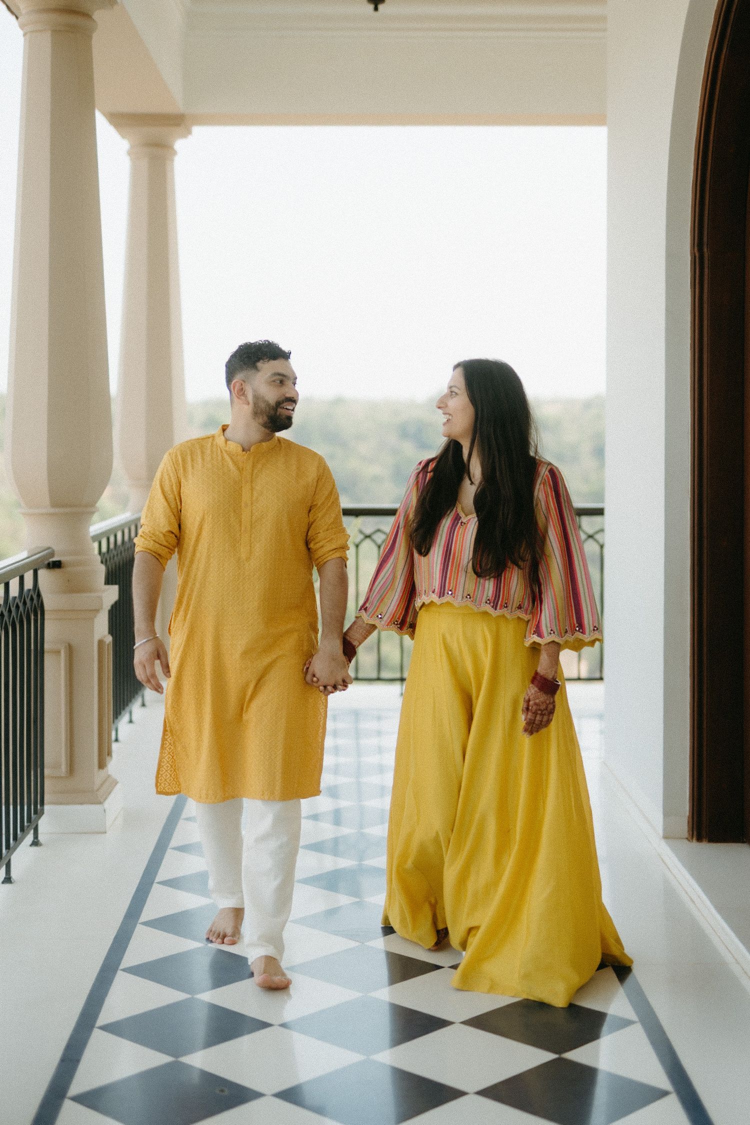 A couple in traditional Indian attire walk together on a black and white checkered balcony wearing yellow and pink outfits.