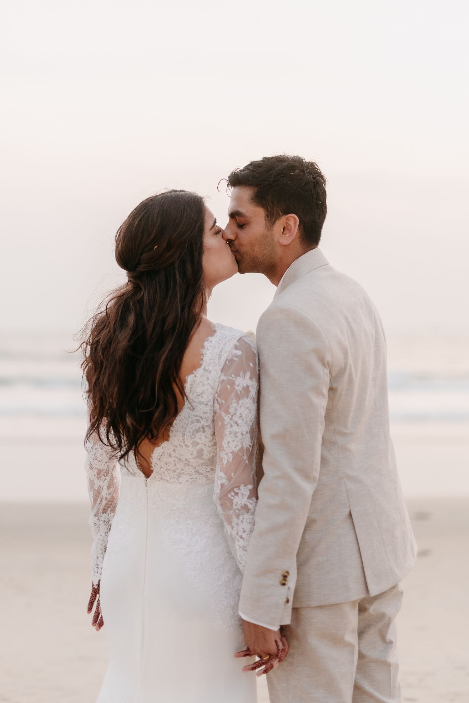 Romantic beach wedding photo of couple kissing at sunset in lace white dress and light beige suit.
