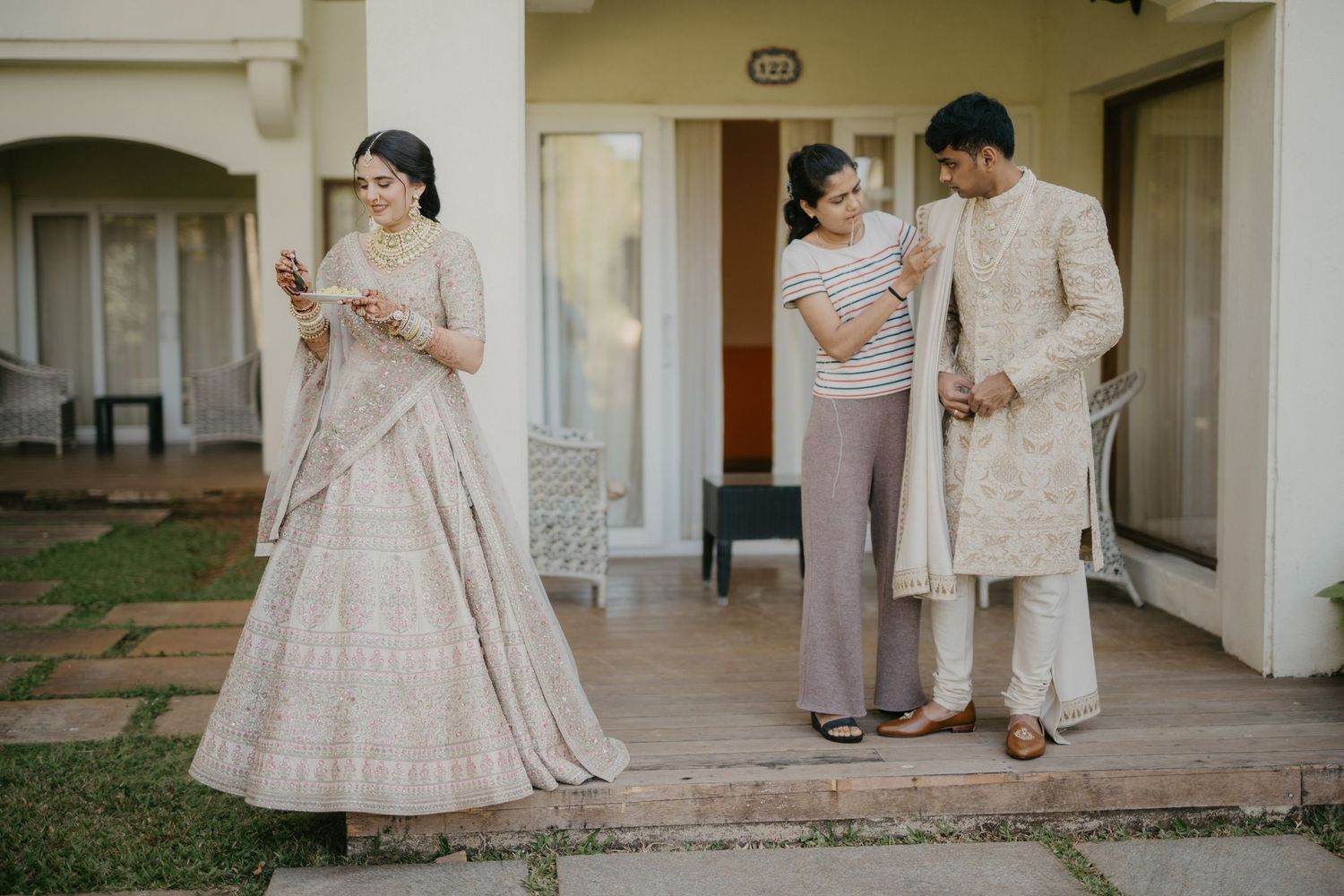 Couple dressed in elegant light-colored traditional Indian attire stands on a porch during a wedding ceremony.