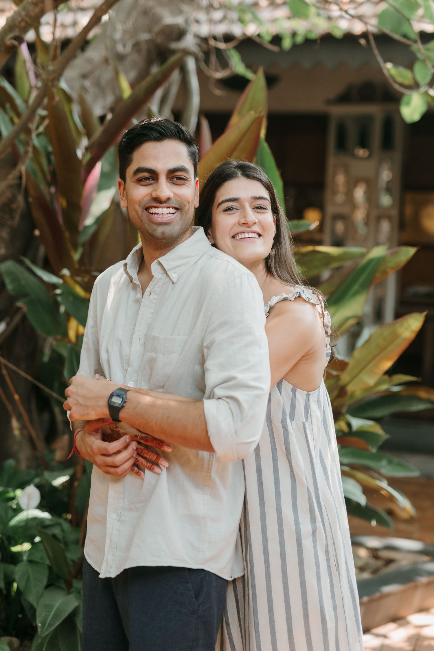 A couple in casual summer outfits smile warmly while posing together against lush green tropical foliage.