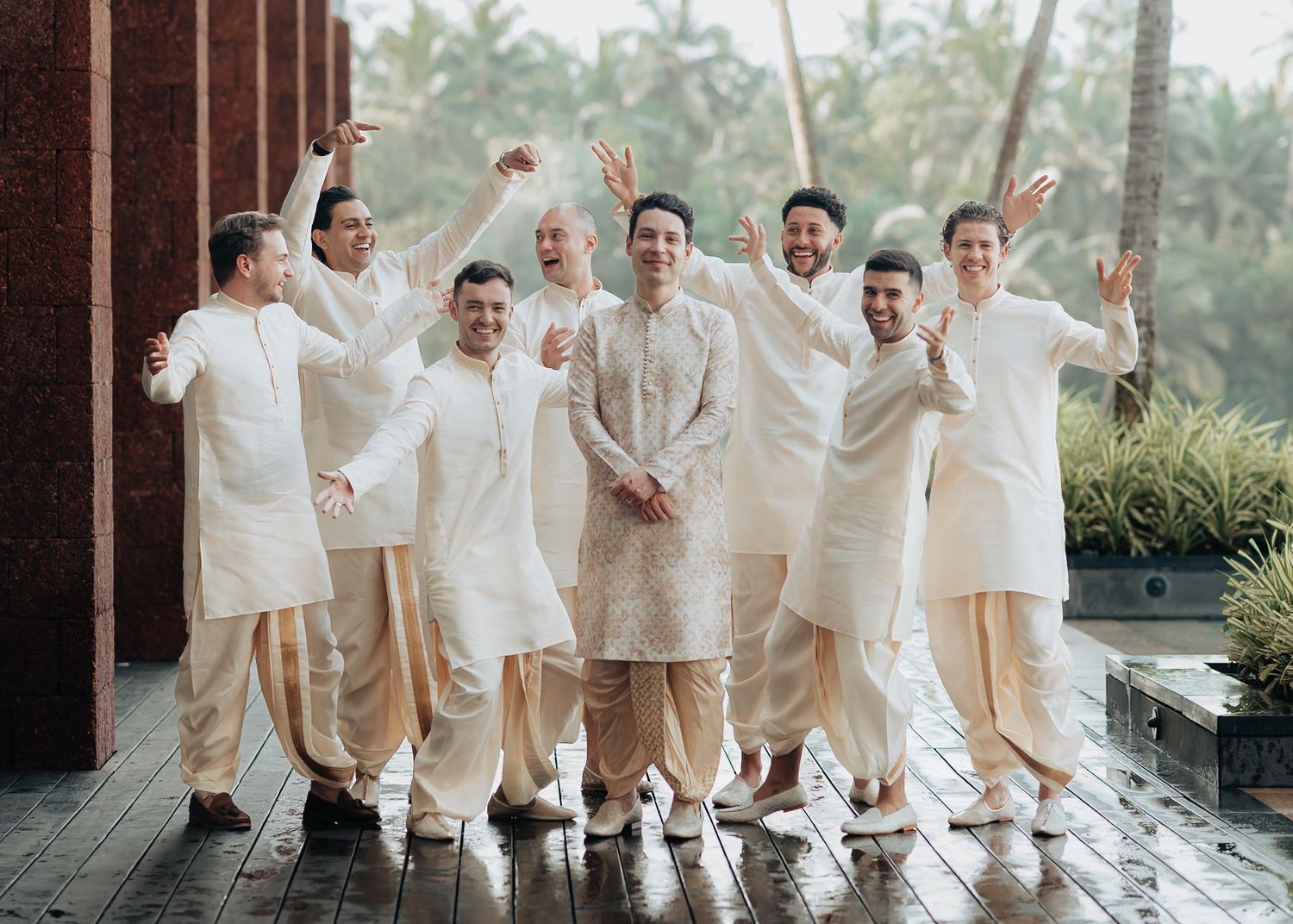 Groomsmen in coordinated white and beige outfits jumping and celebrating outdoors.