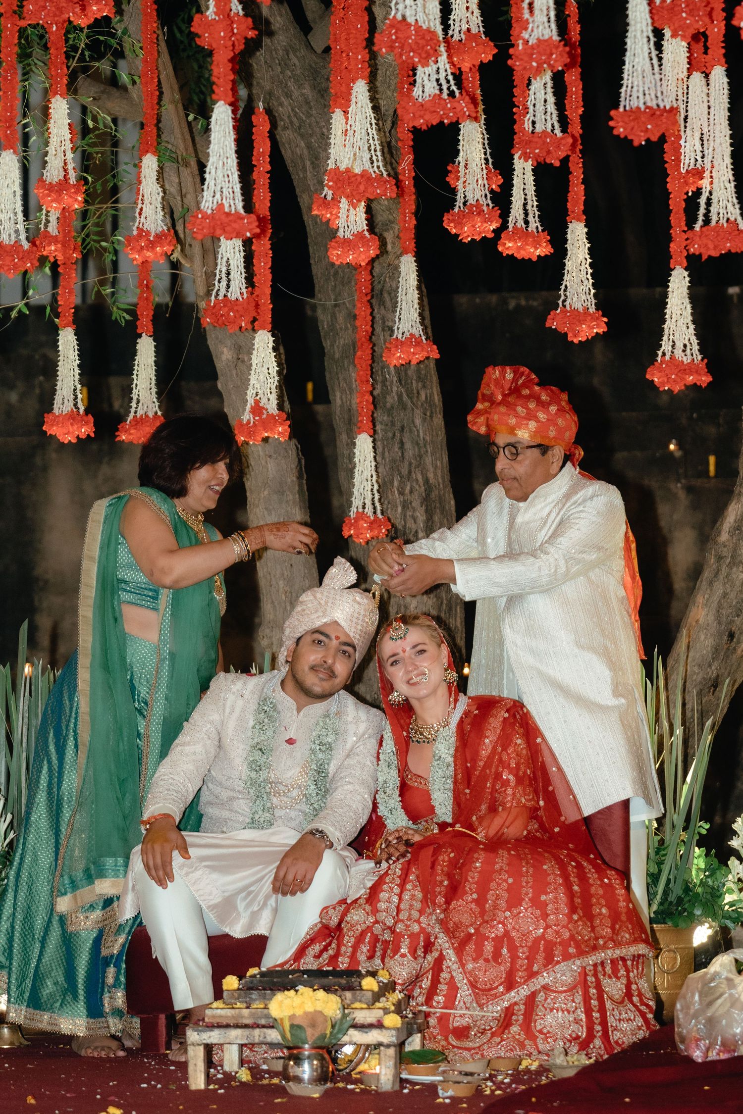 Wedding celebration under decorative red and white hanging flowers with family performing rituals.