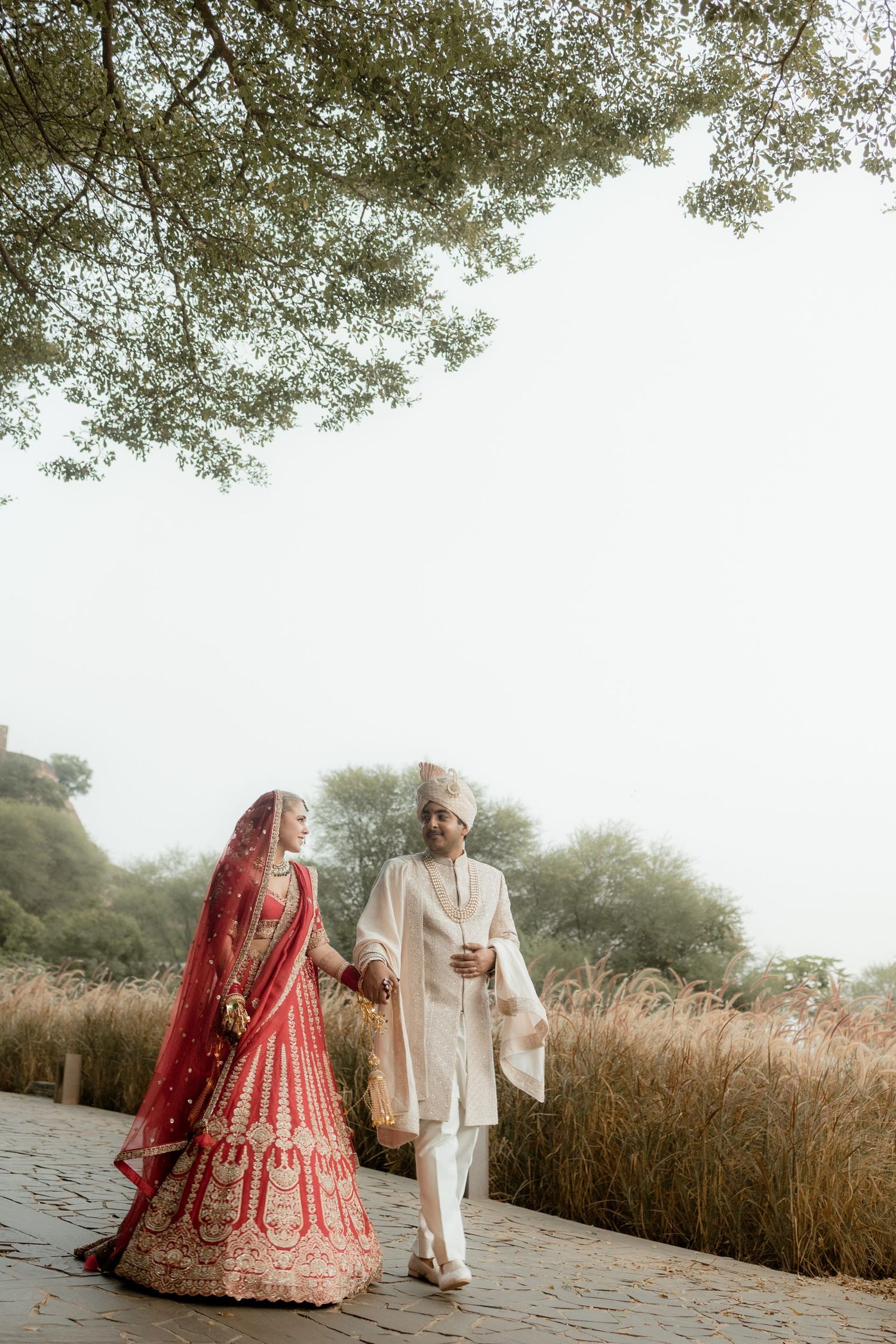 Traditional Indian wedding couple walking through scenic outdoor setting with tall grass.