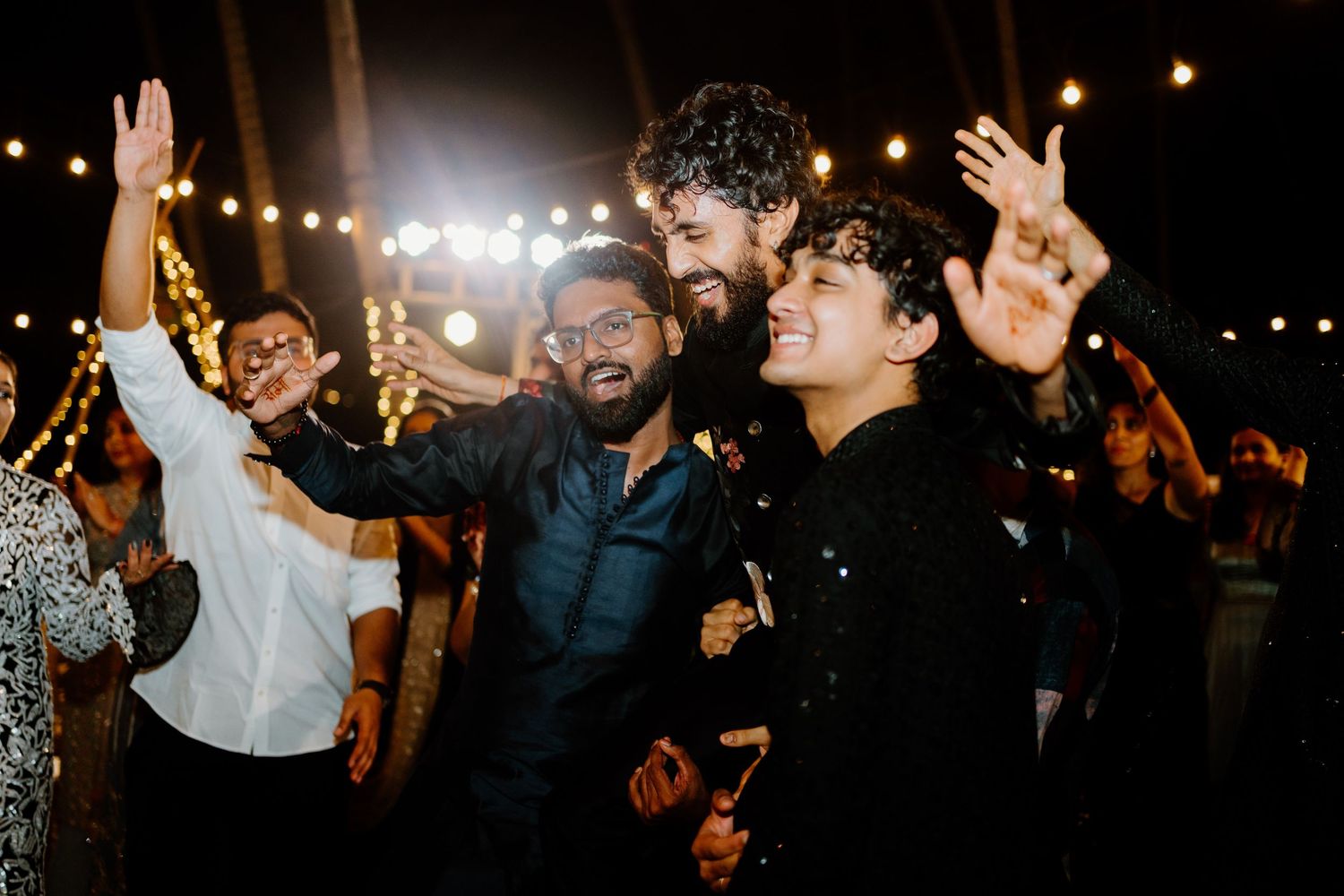 Three joyful men dancing and celebrating at a festive outdoor night party with string lights.