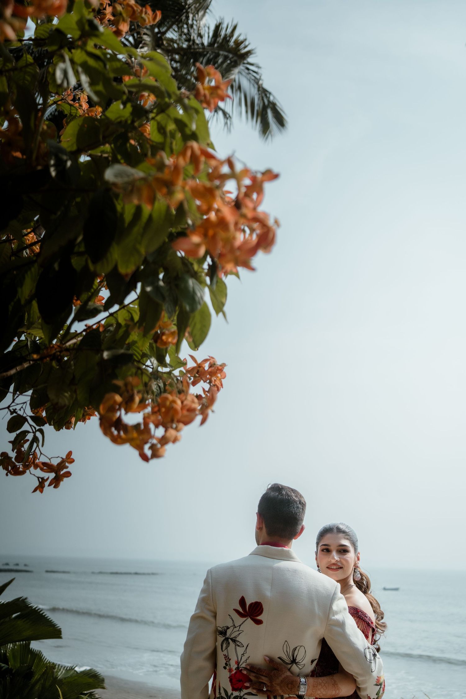 A couple in formal attire sits together on a beach with orange flowering trees and ocean in background.