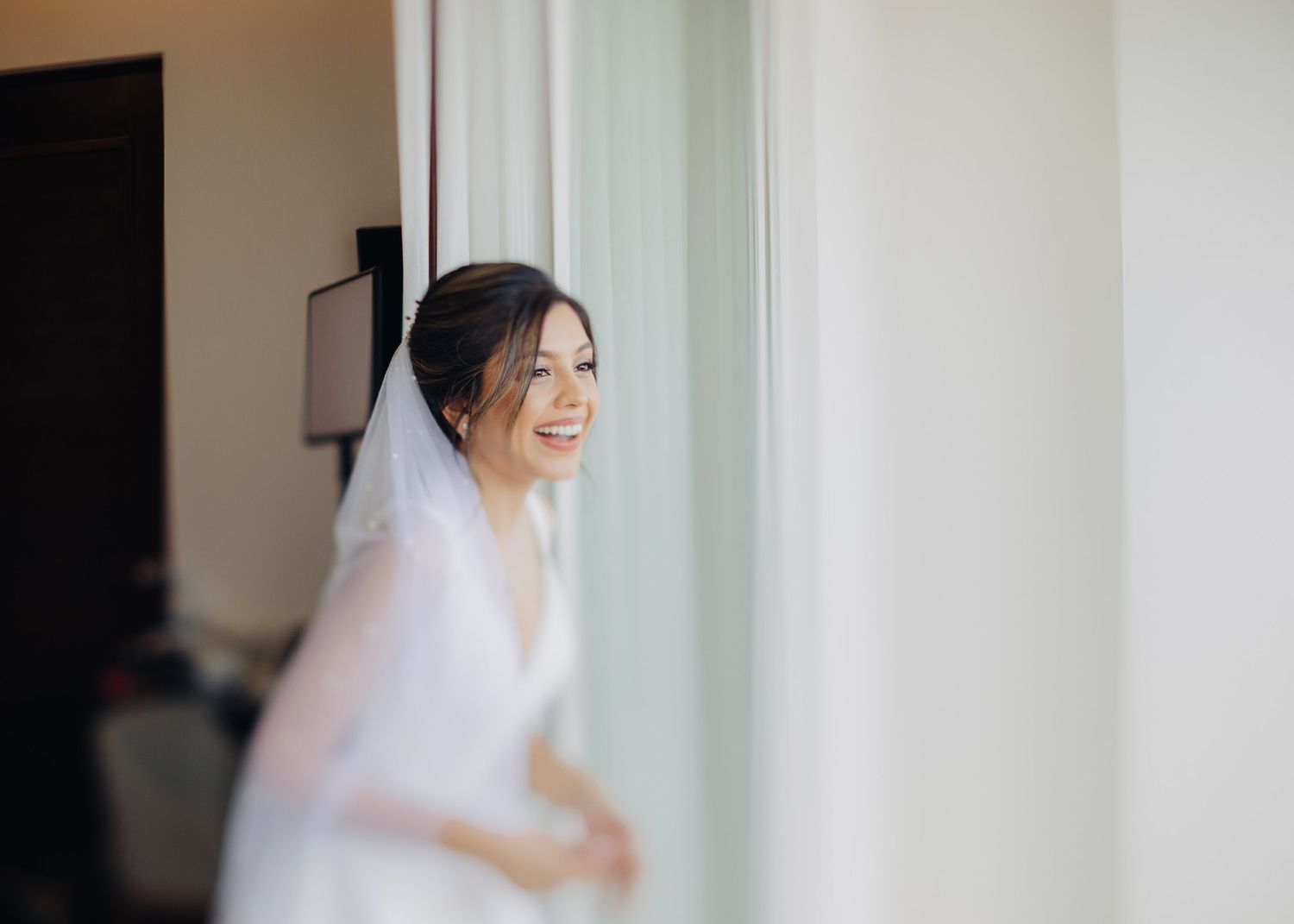 A joyful bride in a white wedding dress and veil stands by a window, looking to the side with a candid smile.