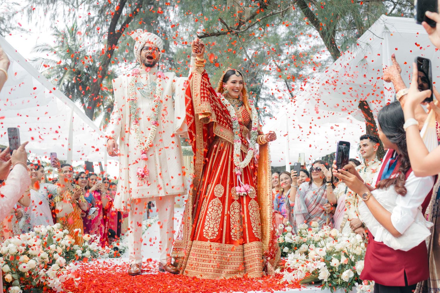 Festive wedding celebration with guests joyfully throwing red confetti in an outdoor garden setting.