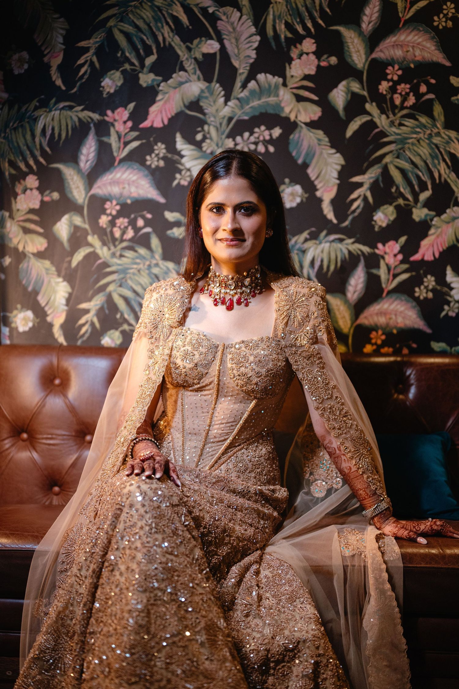 Bride in gold embroidered lehenga with red choker necklace sits elegantly before floral wallpaper backdrop.