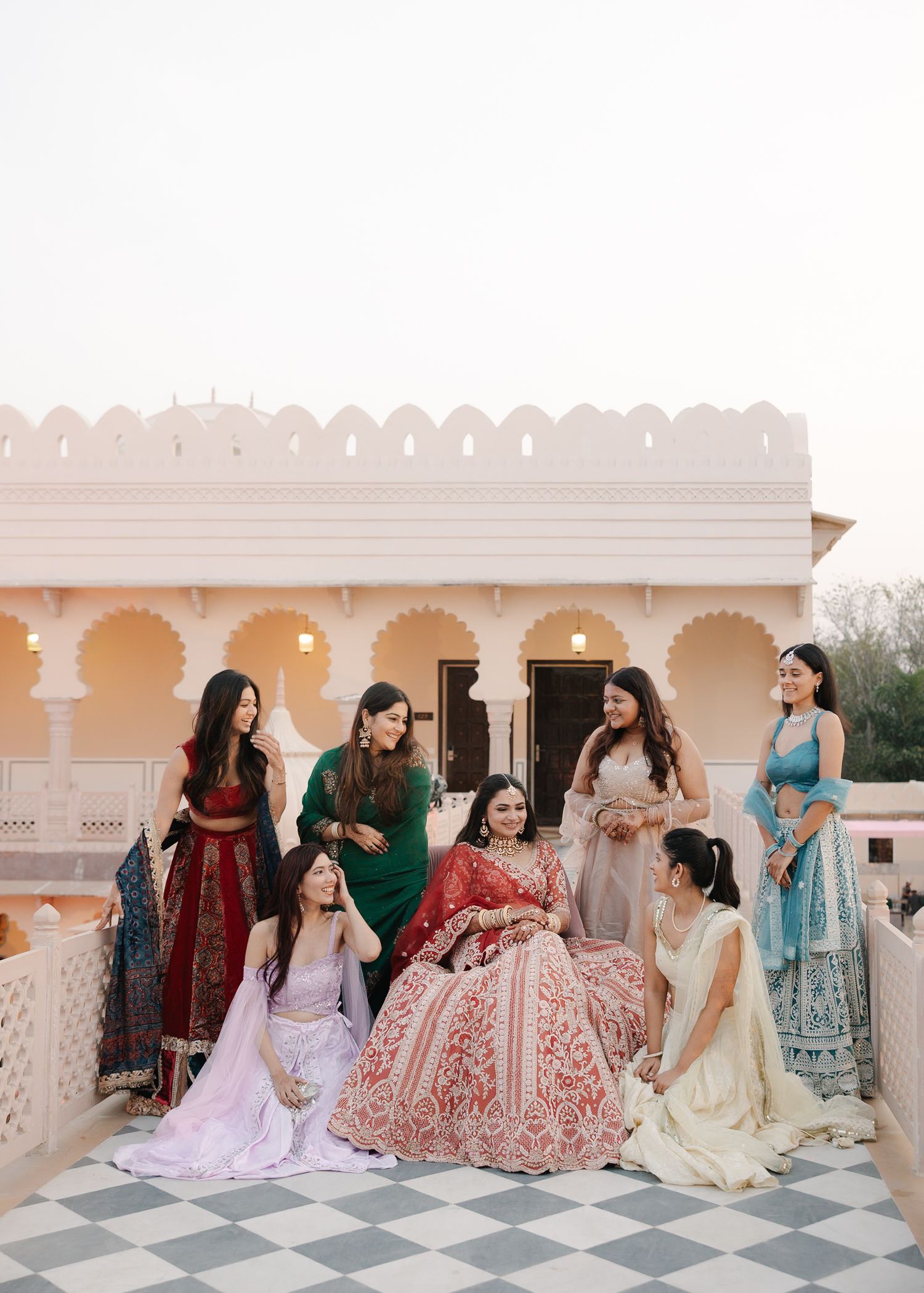Indian bride in red lehenga surrounded by bridesmaids in colorful outfits on palace rooftop.