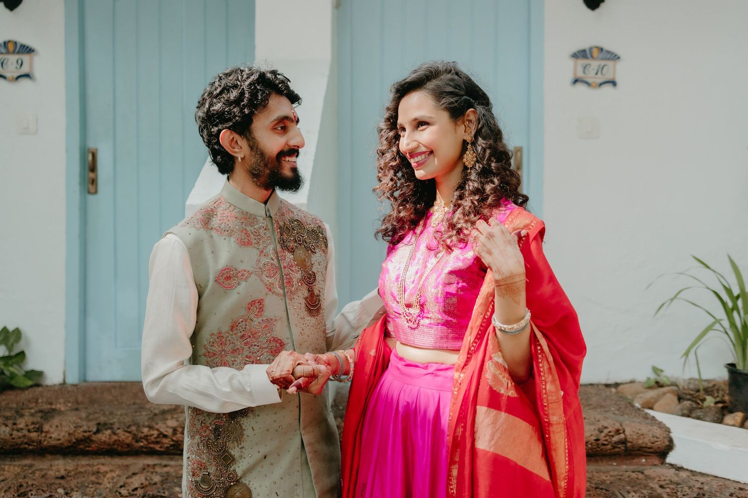 Indian couple in traditional attire smiling at each other outdoors, man in embroidered sherwani, woman in pink lehenga.