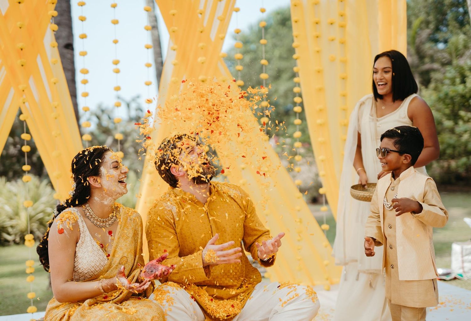 Joyful Indian wedding haldi ceremony with couple in yellow outfits, flower petals thrown by children outdoors.