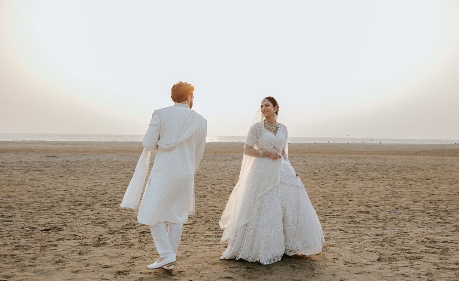 Couple in white wedding attire standing on a sandy beach during golden hour.