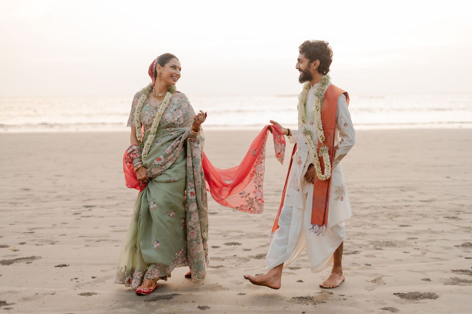 Couple in traditional Indian attire on a beach, woman in green saree, man in white kurta, sharing a joyful moment.