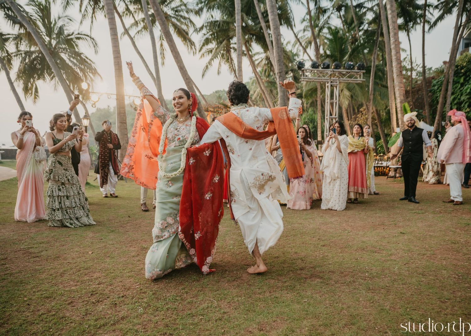 Wedding celebration on a beach with palm trees and guests in traditional attire.