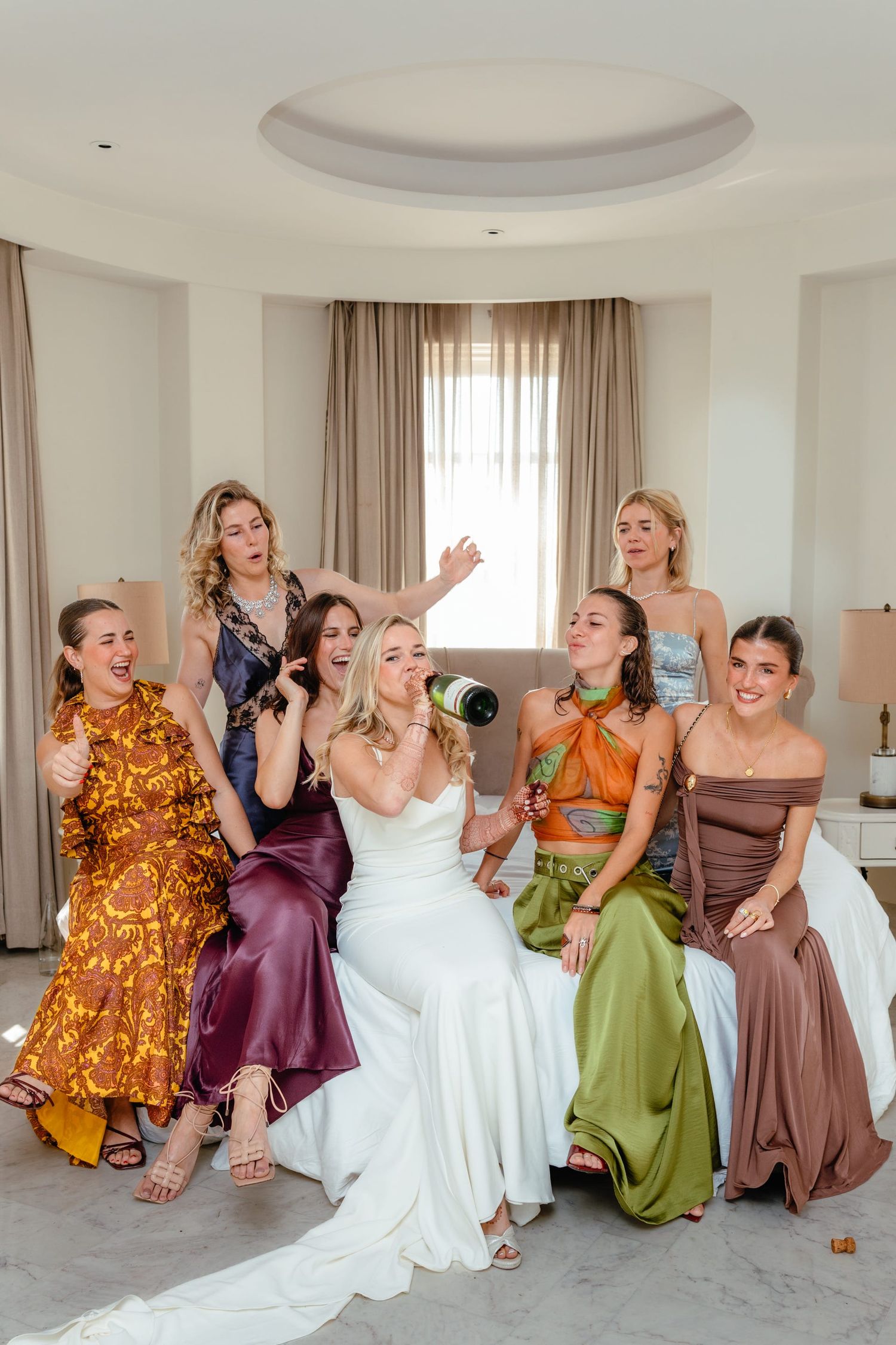 Wedding party laughs together while popping champagne in a sunlit room with elegant decor.