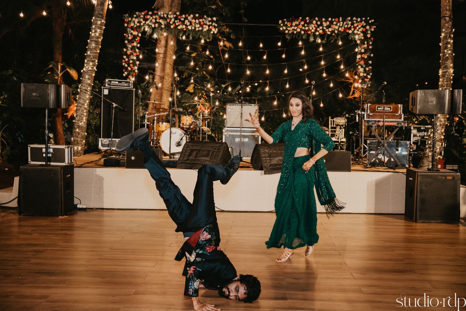 Dancers perform acrobatic breakdancing moves on a wooden dance floor with string lights overhead.