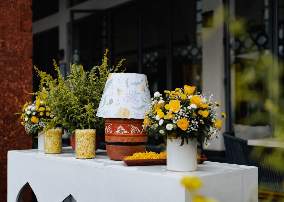 Table display with yellow floral arrangements and decorative lamp against white backdrop.