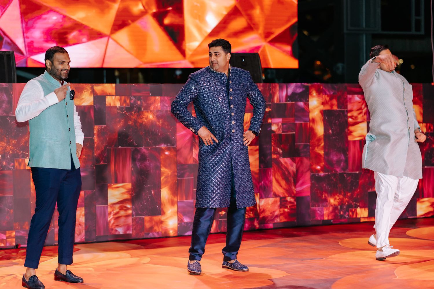 Three men in traditional Indian attire perform on a vibrant stage with red mosaic backdrop at a cultural event.