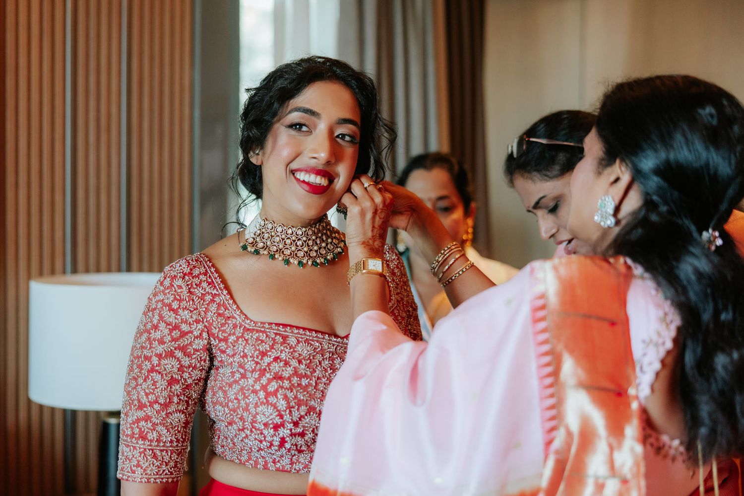 Indian bride in red lehenga smiles as woman helps with gold jewelry during wedding preparation.