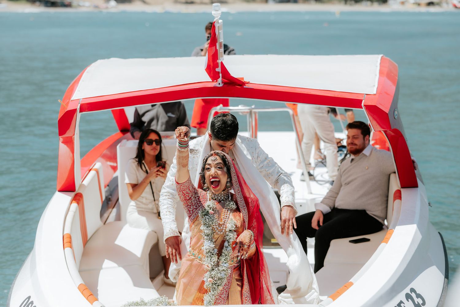 Joyful Indian bride and groom celebrate on a speedboat with guests after wedding ceremony.