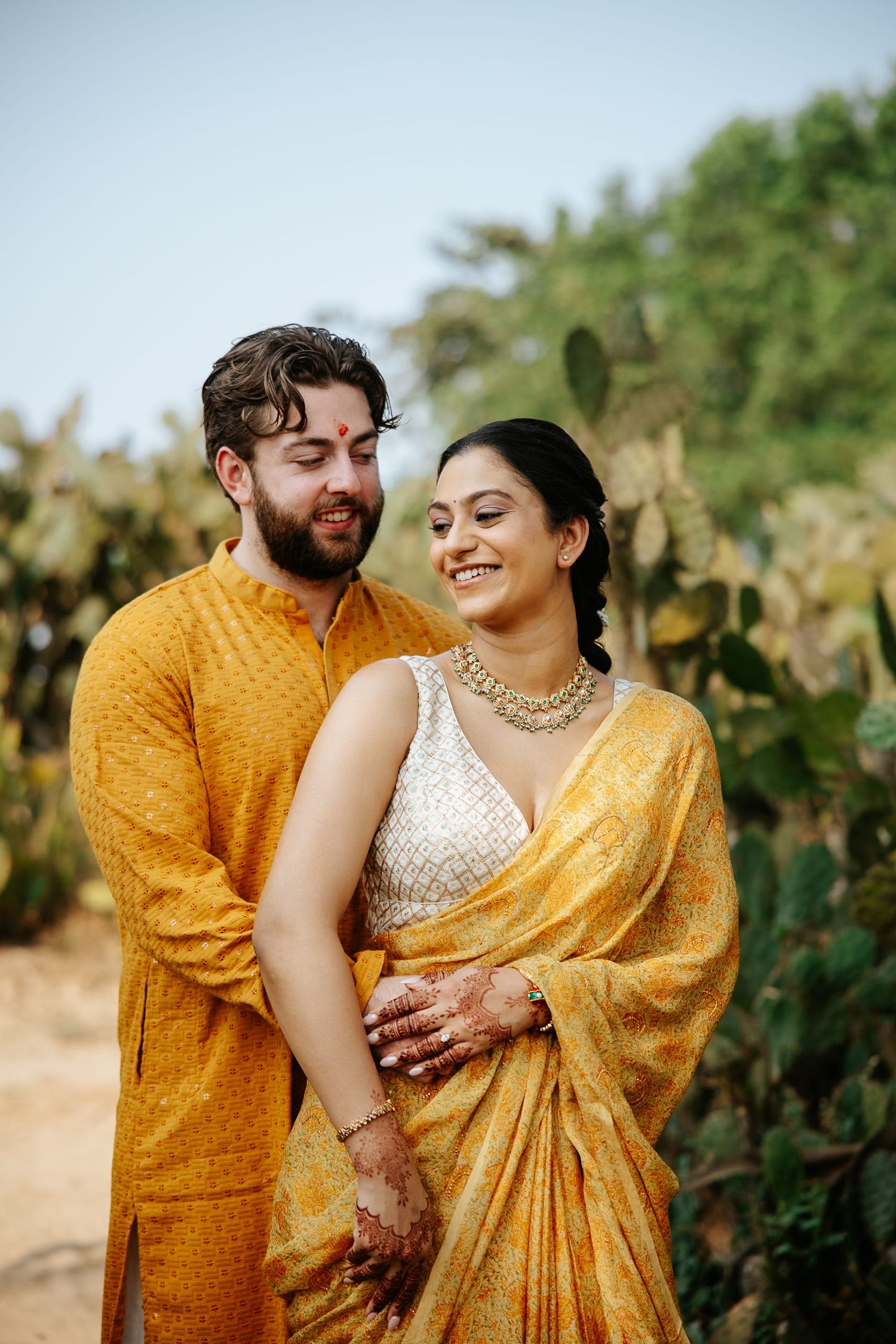 Couple in matching mustard yellow outfits smiling together outdoors, woman in saree with gold jewelry, man in kurta.