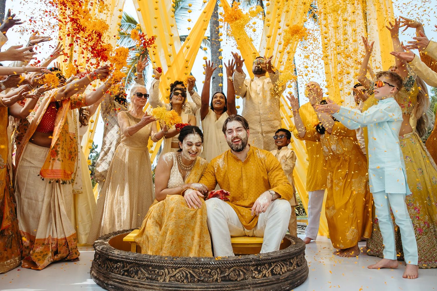 Couple seated in ornate vessel during vibrant Indian Haldi ceremony, surrounded by guests in yellow and gold attire tossing flowers.