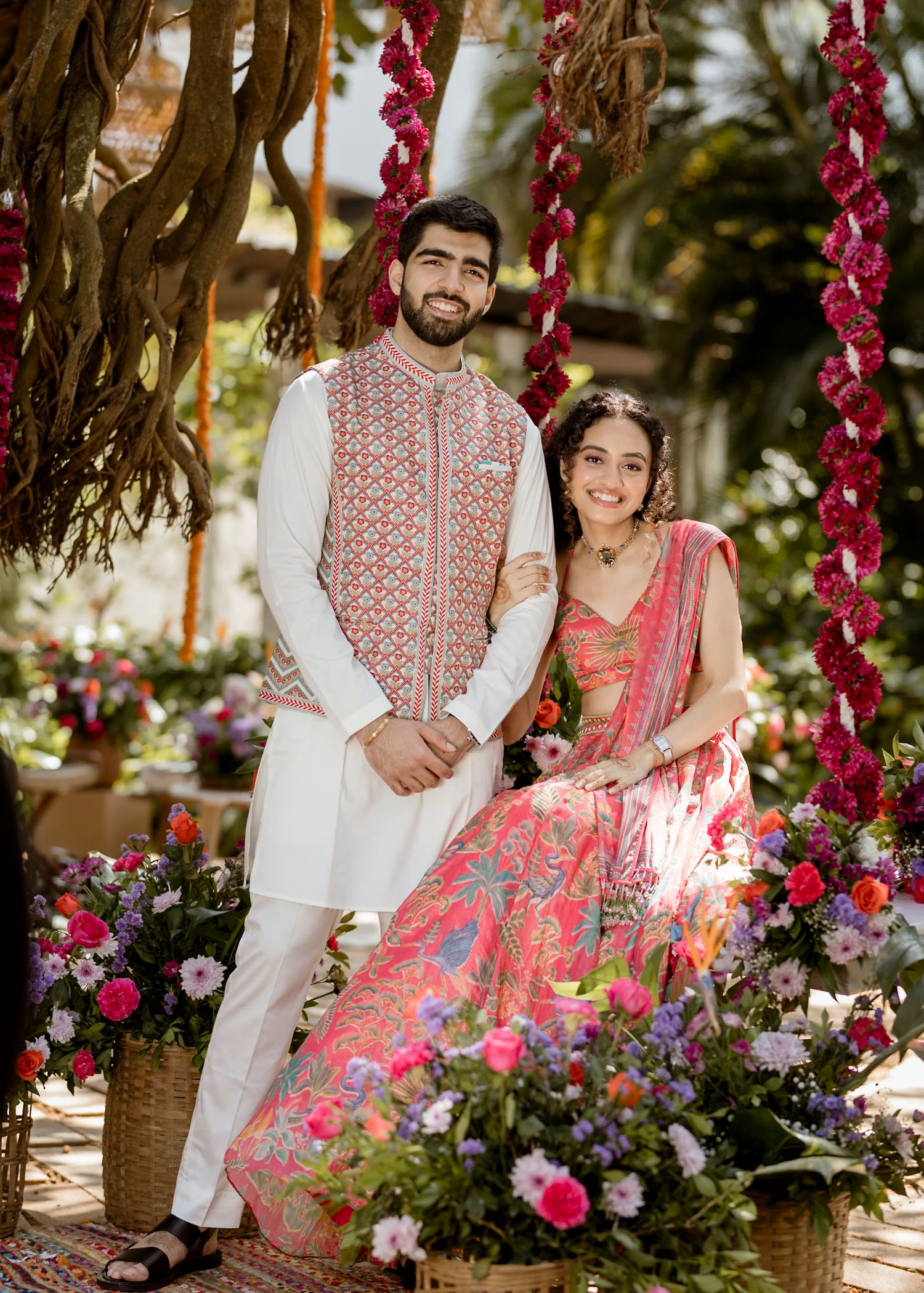 Couple in traditional Indian attire posing among floral decorations and pink garlands.