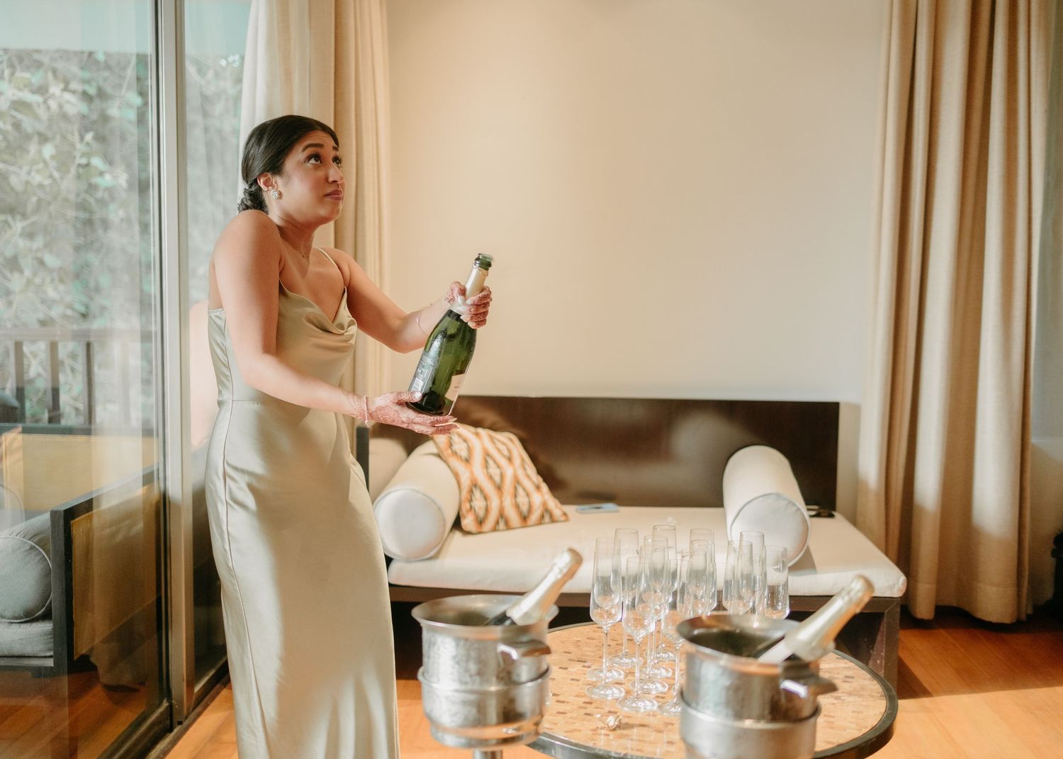 A person in an elegant gown pours champagne into glasses at a stylishly set table with silver accessories.