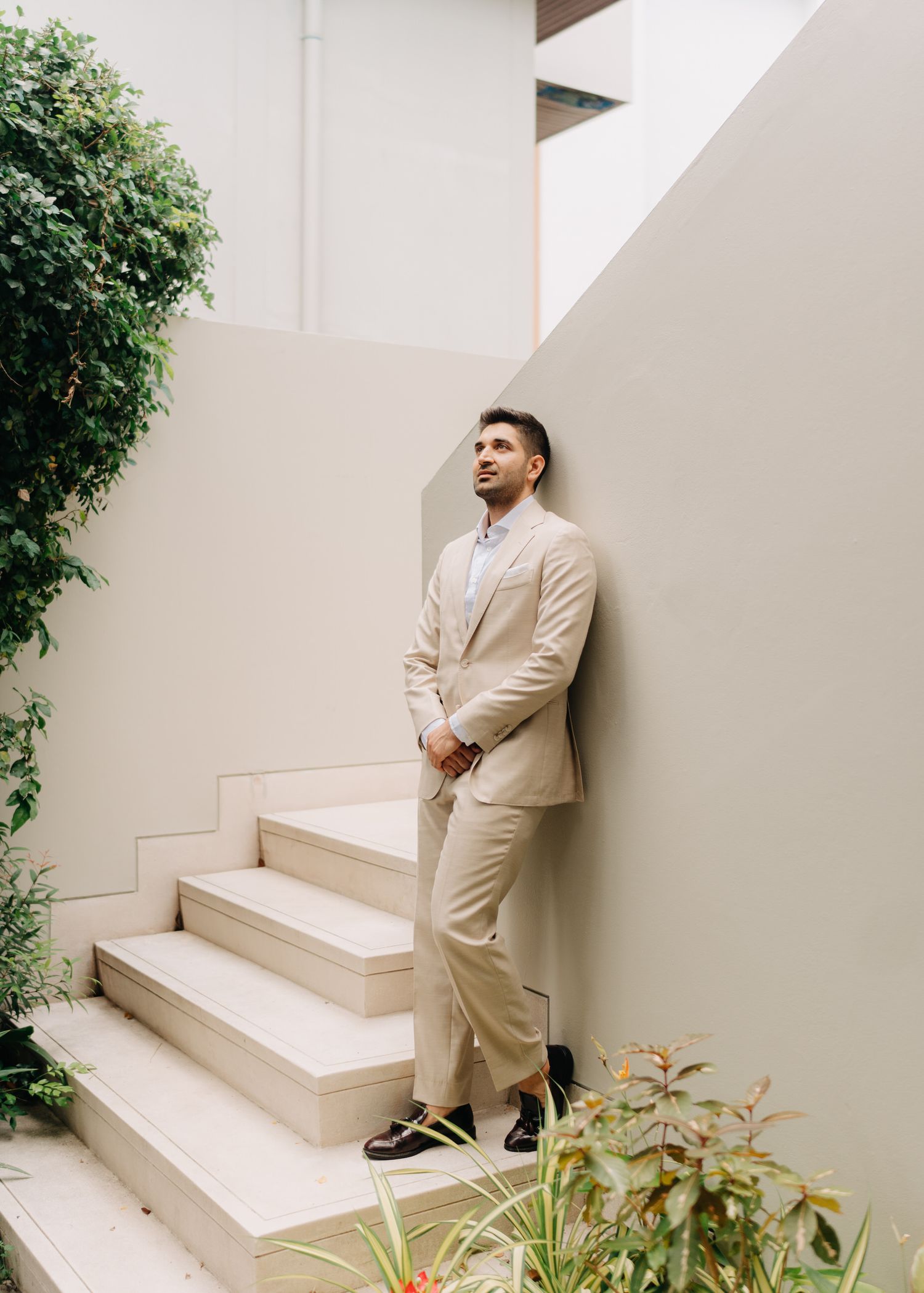 A person wearing a beige outfit poses against a white wall with steps and greenery in a minimalist outdoor setting.