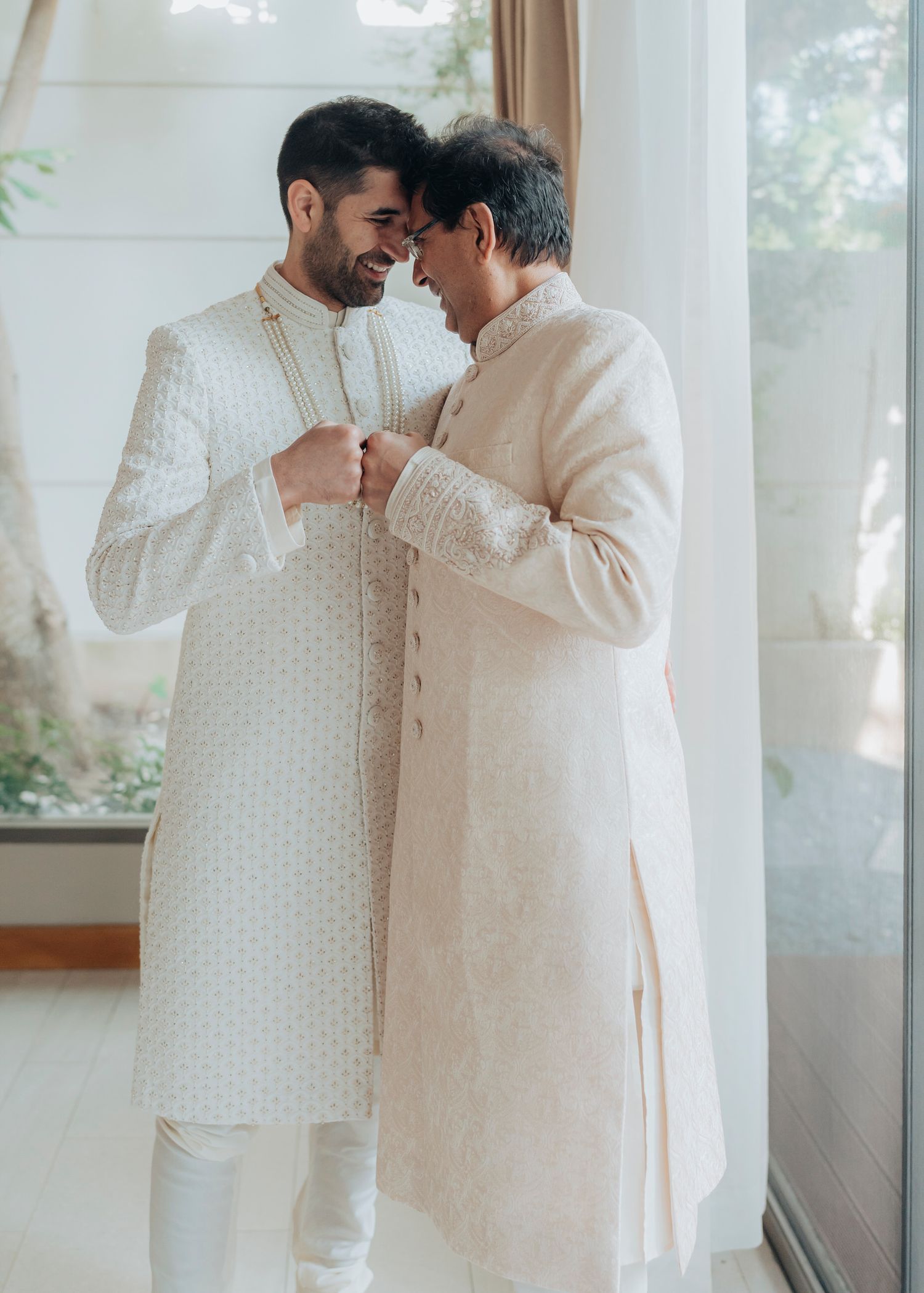 Two people in traditional white Indian wedding attire share an intimate moment by a window.