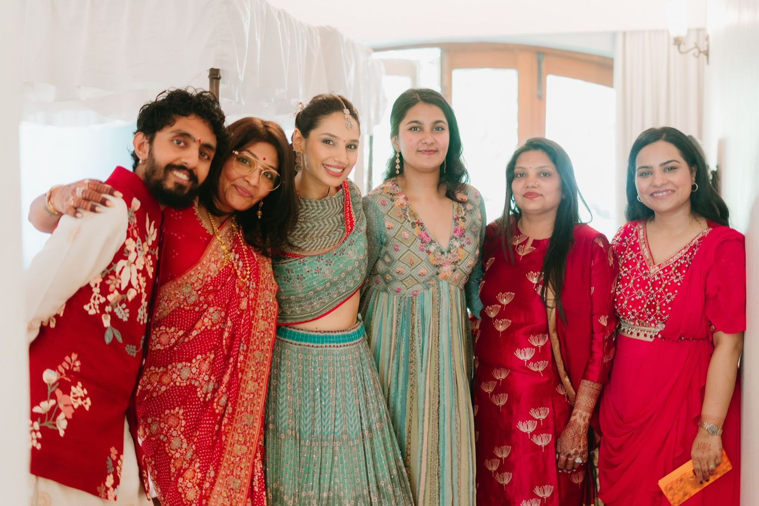 Group of six people in vibrant Indian attire, including red and teal outfits, posing together at a festive celebration.