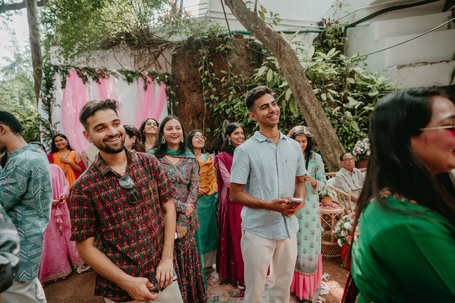 Two men smiling at an outdoor Indian celebration with colorful traditional attire and festive decor in the background.