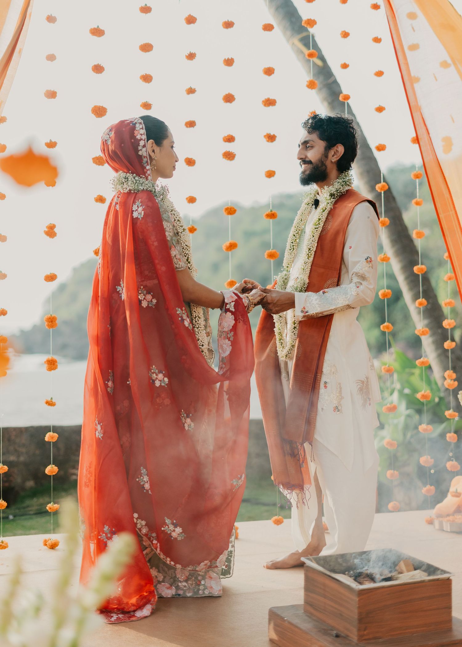 Couple in traditional Indian wedding attire holding hands under marigold garland decorations at an outdoor ceremony.