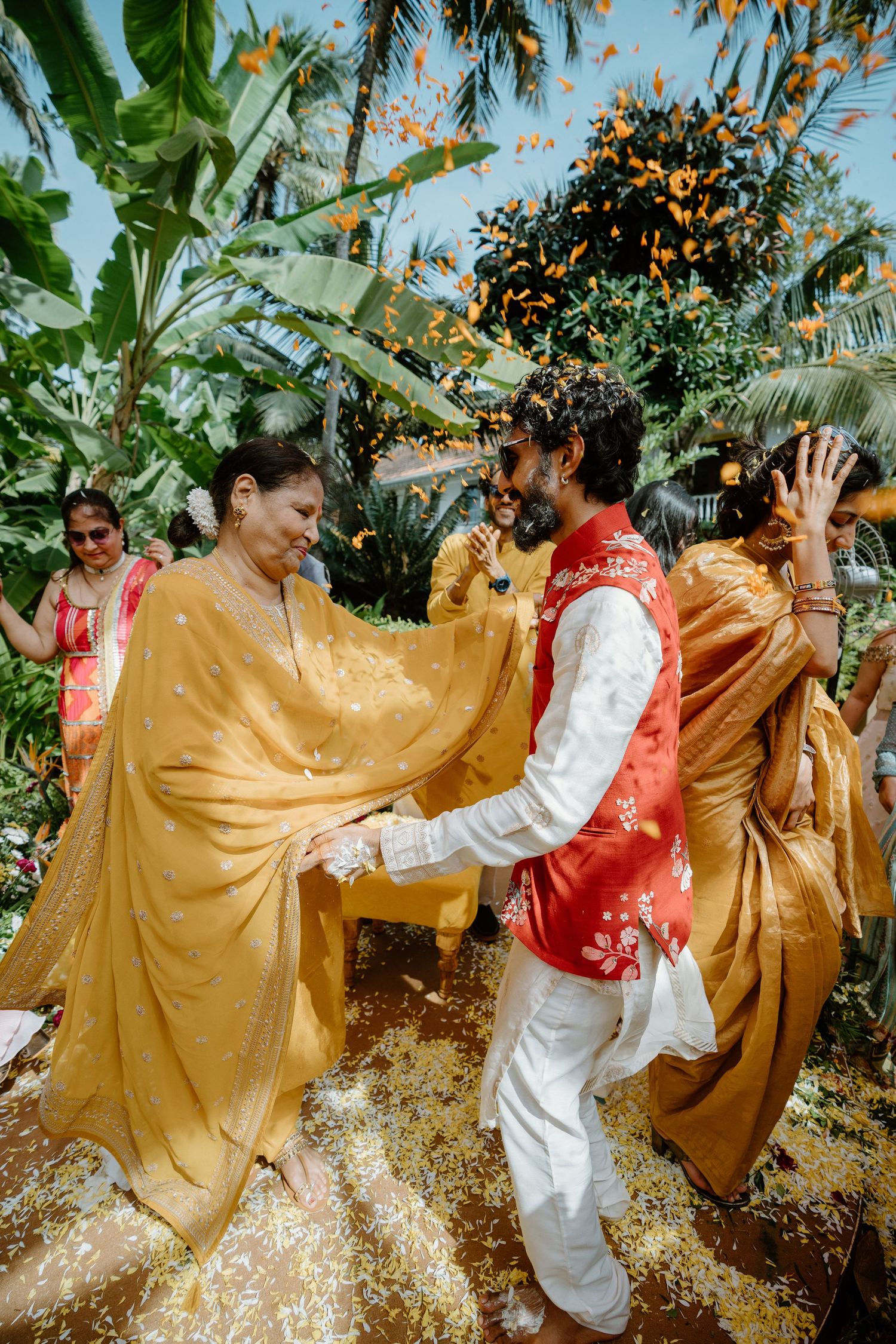Joyful Indian wedding celebration with guests in yellow and red traditional attire, dancing outdoors with marigold petals falling.