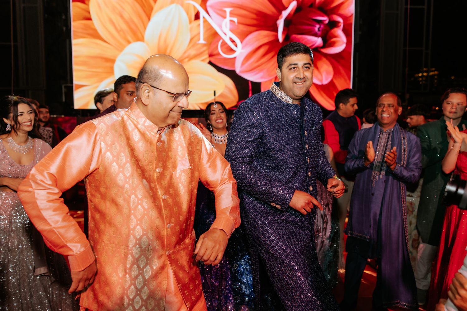 Two men in elegant kurtas share a joyful dance moment at an Indian wedding reception.