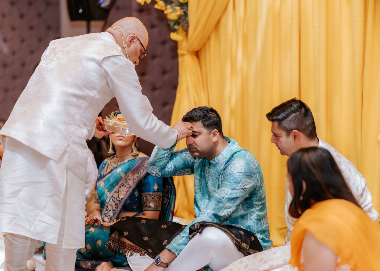 Elder blesses groom during traditional Indian wedding ceremony with bride in colorful sari and yellow drape backdrop.